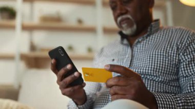 Yaşlı bir Afrikalı Amerikalı adam elinde kredi kartı tutarken akıllı telefonuna odaklanarak evinde rahatça oturuyor. Kişisel alanında meşgul ve rahat görünüyor..