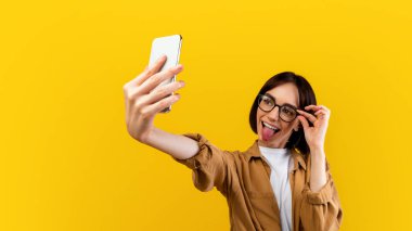 Oyuncu kadın selfie çekiyor, cep telefonu kamerasına dil gösteriyor, gözlüklere dokunuyor, sarı arka planda poz veriyor, stüdyo çekimi yapıyor. Neşeli bayan eğleniyor ve fotoğraf çekiyor.
