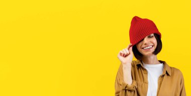 Mutlu genç bir kadın moda mor şapkasını indiriyor, bir gözünü kapatıyor ve gülümsüyor, turuncu arka planda eğleniyor ve oynaşıyor, stüdyoda çekim yapıyor.