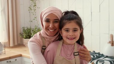 Sıcak ve davetkar bir mutfakta tesettürlü bir anne kızını sevgiyle kucaklıyor. Gülümserler, birlikte yemek pişirirken güçlü bağlarını sergilerler, ailecek geçirdikleri zamanın tadını çıkarırlar..