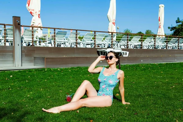 さわやかな夏のカクテルを飲んだり 草の上に座って水着で魅力的な若い女性 — ストック写真