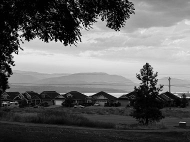 Monochromatic houses line a street, framed by dark trees, with a serene lake and layered mountains fading into the cloudy horizon.