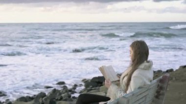 Genç bir kız akşam gün batımında bir kitap tarafından deniz okuma bir bankta oturur. gün batımında sahilde okuma 4 k. A çocuk. Deniz kenarında bir kız rahatlatıcı sahil şeridi.