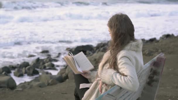 En ung flicka sitter på en bänk vid den hav läser en bok vid solnedgången på kvällen dagen. 4 k. A barn läsning på stranden vid solnedgången. En flicka som är avkopplande vid havet, kusten. — Stockvideo