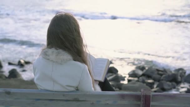 En ung flicka sitter på en bänk vid den hav läser en bok vid solnedgången på kvällen dagen. 4 k. A barn läsning på stranden vid solnedgången. En flicka som är avkopplande vid havet, kusten. — Stockvideo