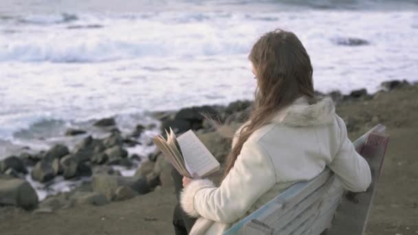 En ung flicka sitter på en bänk vid den hav läser en bok vid solnedgången på kvällen dagen. 4 k. A barn läsning på stranden vid solnedgången. En flicka som är avkopplande vid havet, kusten. — Stockvideo