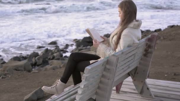 Uma menina senta-se em um banco junto ao mar lê um livro ao pôr-do-sol do dia da noite. 4K. Uma criança a ler na praia ao pôr-do-sol. Uma menina relaxante à beira-mar, litoral . — Vídeo de Stock