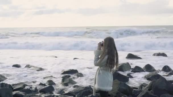 Fotógrafo menina na costa do mar tempestuoso faz fotos. Fotógrafo viajante turístico fazendo fotos oceano. Mar tempestuoso com nuvens de tempestade. Instantâneos de uma viagem ao mar.4k.30fps . — Vídeo de Stock