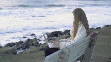 Genç güzel kız güneşin deniz kenarında hayran bir dizüstü bilgisayar baskılar metin 4 k, 30 kare/sn ile bir bankta oturur