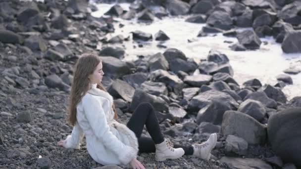 一个年轻女孩坐在海滩上欣赏美丽的日落 并向海里投掷鹅卵石 — 图库视频影像