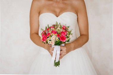 Gelin buketi, çıplak omuzlu, beyaz gelinlik giymiş gelinin ellerinde. Buket küçük pembe güller ve beyaz kurdeleyle tamamlanmış diğer küçük sarı-beyaz çiçeklerden oluşur. Bukete odaklan..