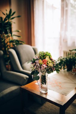 Güllerden oluşan buket, diğer çiçek türleri, dekoratif yapraklar ve dantelli beyaz bir kurdele ile tamamlanmış. Buket, eski bir oturma odası kahve masasının üzerinde duran cam bir vazoya konur..