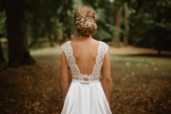 Bride in park facing back in white wedding dress with lace and bare back. Hairs are arranged in braided bun, complemented by tiny flowers. Focus on bride's back, background slightly out of focus.