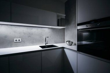 Minimalist tasarımlı modern beyaz mutfak, geceleri, ışık şeridi açık, modern fırın, granit lavabo, duvar prizleri ve cam, beton, alüminyum ve paslanmaz çelik gibi özel malzemeler.
