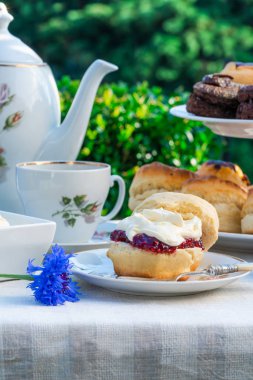 Afternoon tea with cakes and traditional English scones with strawberry jam and clotted cream set up on a table in the garden. Outdoor dining. clipart
