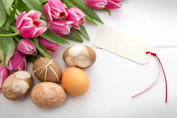 Påskkort med bukett rosa tulpaner och färgglada ägg på vitt trä . — Stockfoto