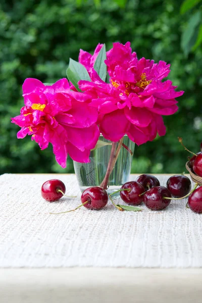 Cerezas grandes y peonía roja en vaso de agua aislado en verde. —  Fotos de Stock