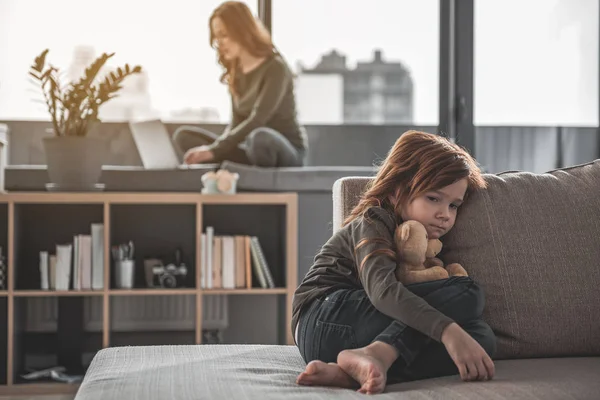 Flicka sitter ensam på soffan medan mor använder laptop — Stockfoto
