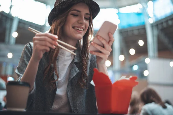 Mulher sorridente está usando o telefone enquanto come — Fotografia de Stock