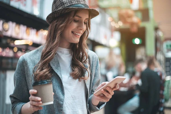 Улыбающаяся девушка с кофе использует смартфон — стоковое фото