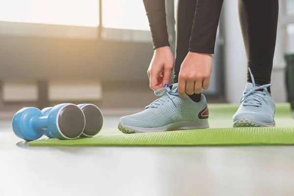 Close up of woman hands and legs standing on green sport mat. She is leaning close to blue dumbbells holding sneakers laces in fingers before exercising