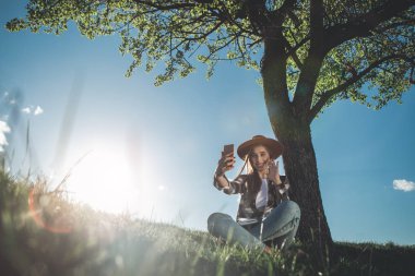 Merhaba. Tam uzunlukta yaz yeşil çayır üzerinde oturan güzel genç kadın. O mutluluk ve cep telefonu tutan ve el sallayarak sevinç dolu. Kopya alanı sol tarafında