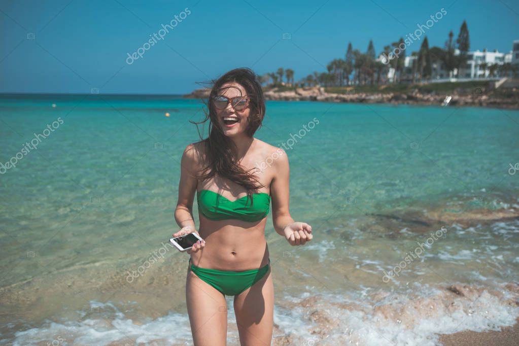 It is a great place. Cheerful slim girl is having fun at the beach. She is holding mobile phone and laughing. Picturesque sea landscape on background 
