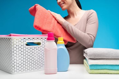 Deterjanlar, clothesbasket ve katlanmış kumaş beyaz masanın üzerinde yatan kapatın. Gülümseyen ev kadını arkasındaki turuncu yumuşatılmış havlu dokunuyor 