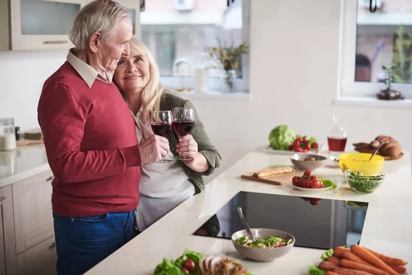Loving elderly couple standing by cooking board full of dishes. They are bonding to each other with glasses of red wine in hands with smile and joy