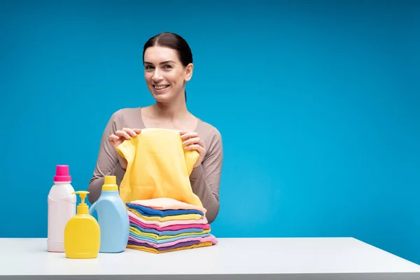 Mulher alegre mostrando roupas limpas à mesa com detergentes — Fotografia de Stock