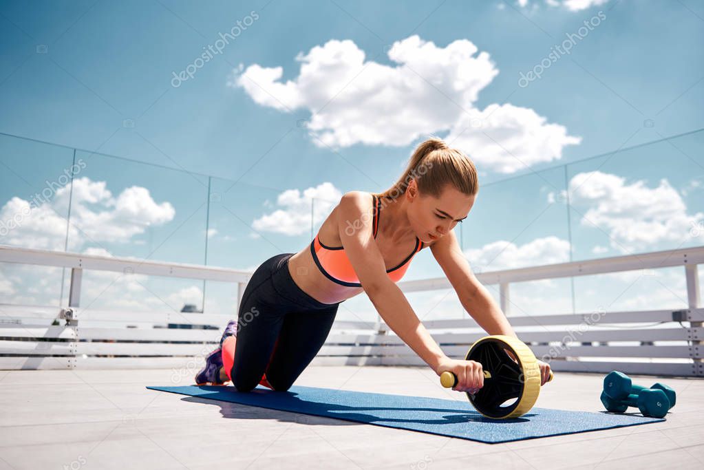 Strained woman is exercising with abs roller on balcony