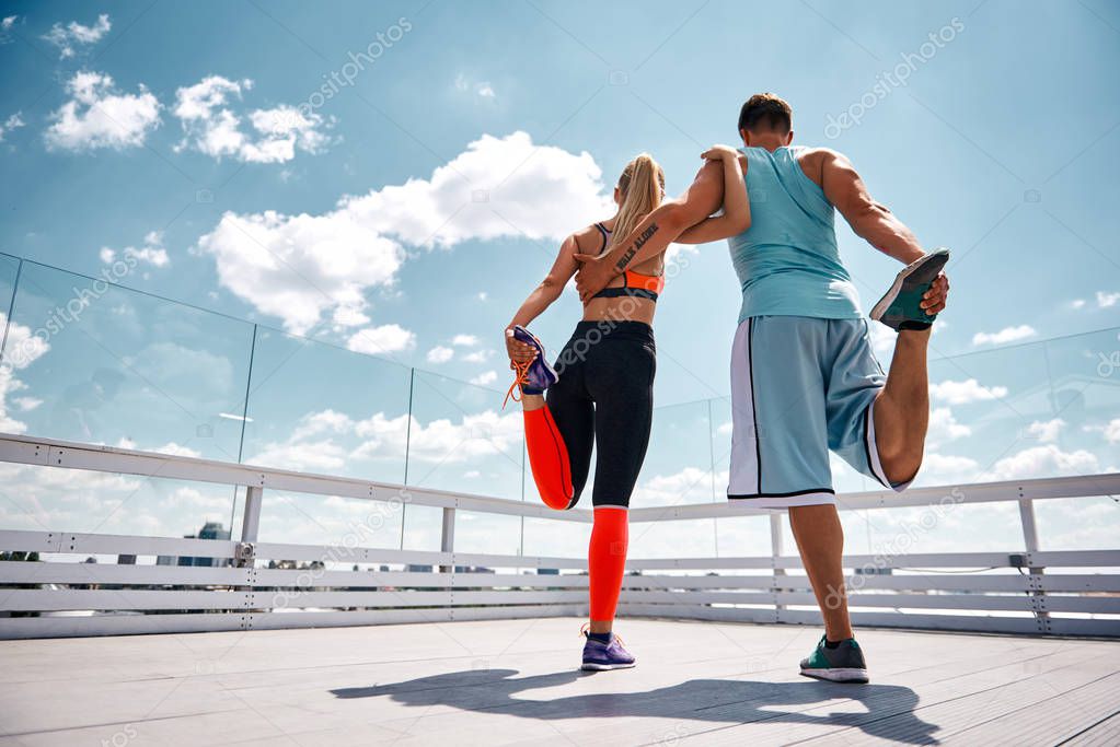 Shapely man and woman are exercising jointly on top of high building. They are standing on one leg and bending other. Partners are supporting each other by embracing while focus is on back