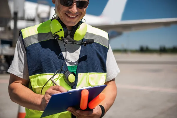 Alegre trabajador del aeropuerto mirando portapapeles y escribir información — Foto de Stock