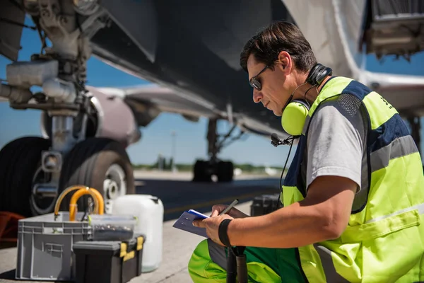 Airport worker squatting near aircraft and writing on clipboard