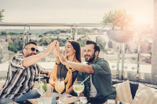 Alegre amigos sentados a la mesa con cócteles y dando cinco altos — Foto de Stock