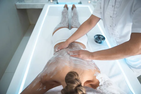 Jovem deitada na mesa de massagem e tendo procedimento de spa com barro branco — Fotografia de Stock