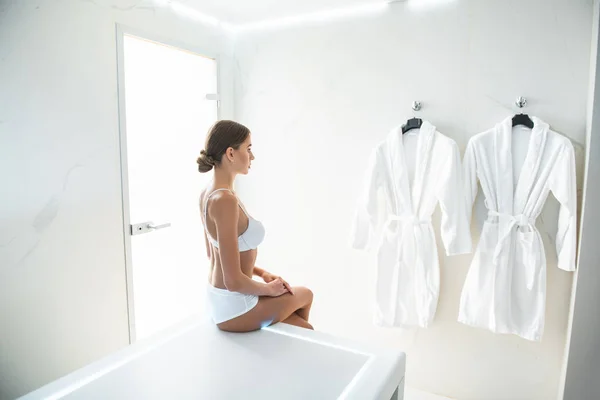 Attractive young lady in white lingerie looking at soft bathrobes on the wall — Stock Photo, Image