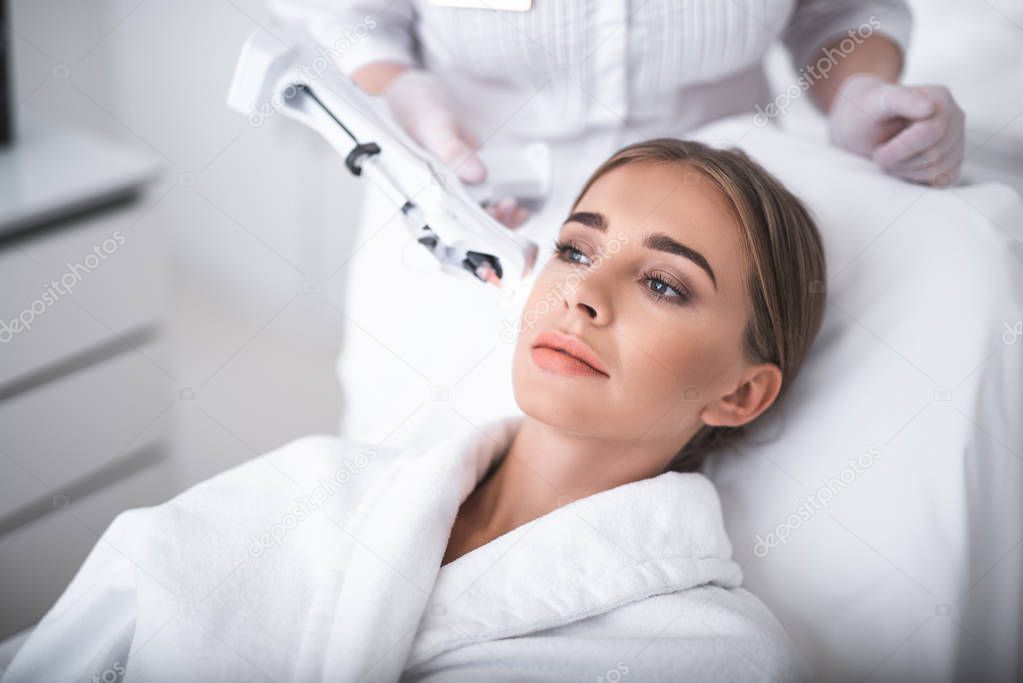 Using syringe gun. Lovely girl in white soft bathrobe having skincare procedure. Cosmetologist hands with needle holder near lady face