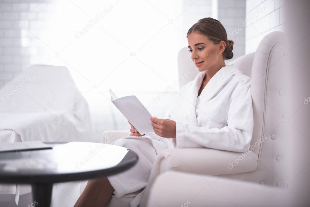 Lovely smiling girl reading at beauty salon while sitting in comfortable armchair