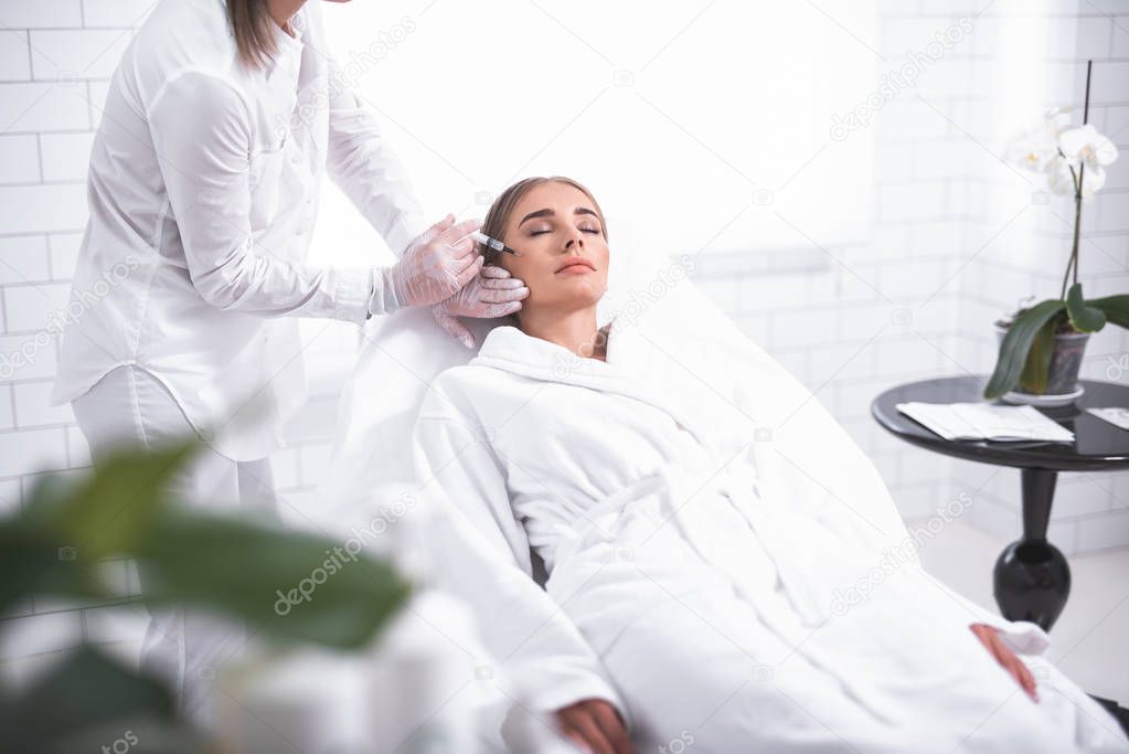 Attractive girl with closed eyes at beauty salon and cosmetologist with syringe