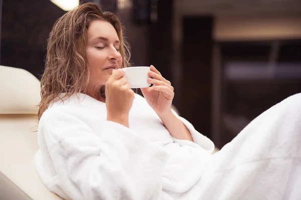 Senhora atraente em roupão de banho branco segurando xícara de bebida quente — Fotografia de Stock