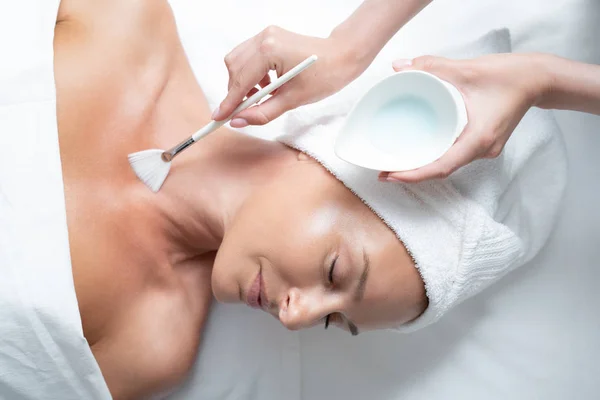 Cosmetologist aplicando máscara para a pele da senhora usando escova e tigela com argila — Fotografia de Stock