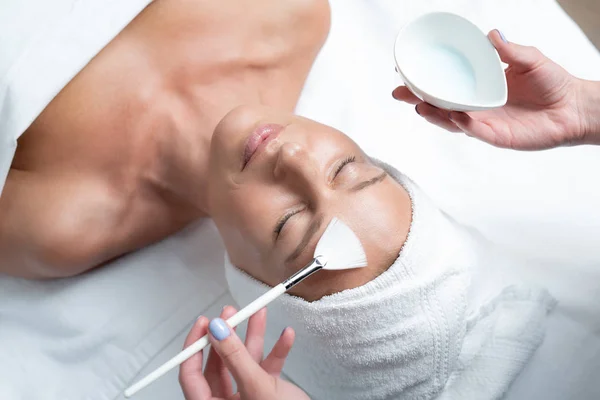 Mãos de esteticista aplicando máscara na testa da senhora usando escova cosmética — Fotografia de Stock