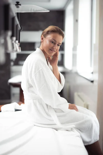 Charmante Frau mittleren Alters im Bademantel sitzt auf Massagetisch im Wellness-Salon — Stockfoto