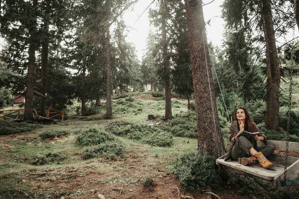 Charmig tjej håller anteckningsbok medan du sitter på trä swing — Stockfoto