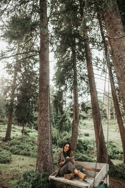 Красивая девушка делает заметки о путешествии, сидя на деревянных качелях — стоковое фото