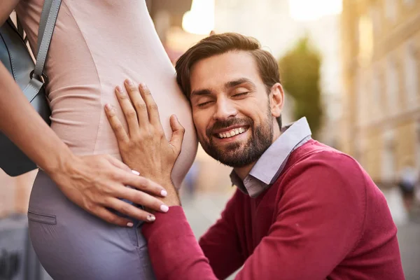 Glücklicher bärtiger Mann, der auf den Bauch seiner schwangeren Frau hört — Stockfoto