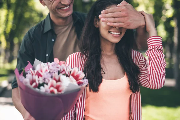 Jovem fechando os olhos de sua namorada e segurando flores para ela — Fotografia de Stock