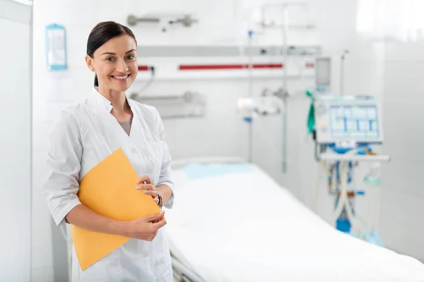 Χαρούμενα νοσοκόμα σε λευκή εργαστήριο παλτό ποζάρει στο δωμάτιο του νοσοκομείου — Φωτογραφία Αρχείου