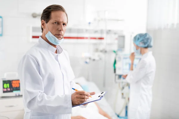 Doctor sosteniendo portapapeles y bolígrafo mientras está de pie en la habitación del hospital — Foto de Stock